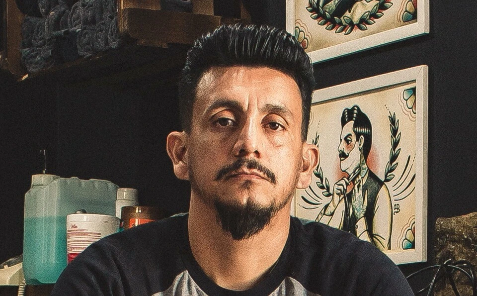 A man with a mustache and goatee, dark pompadour hair, and a dark t-shirt looks at the camera, with framed pictures and bottles in the background.